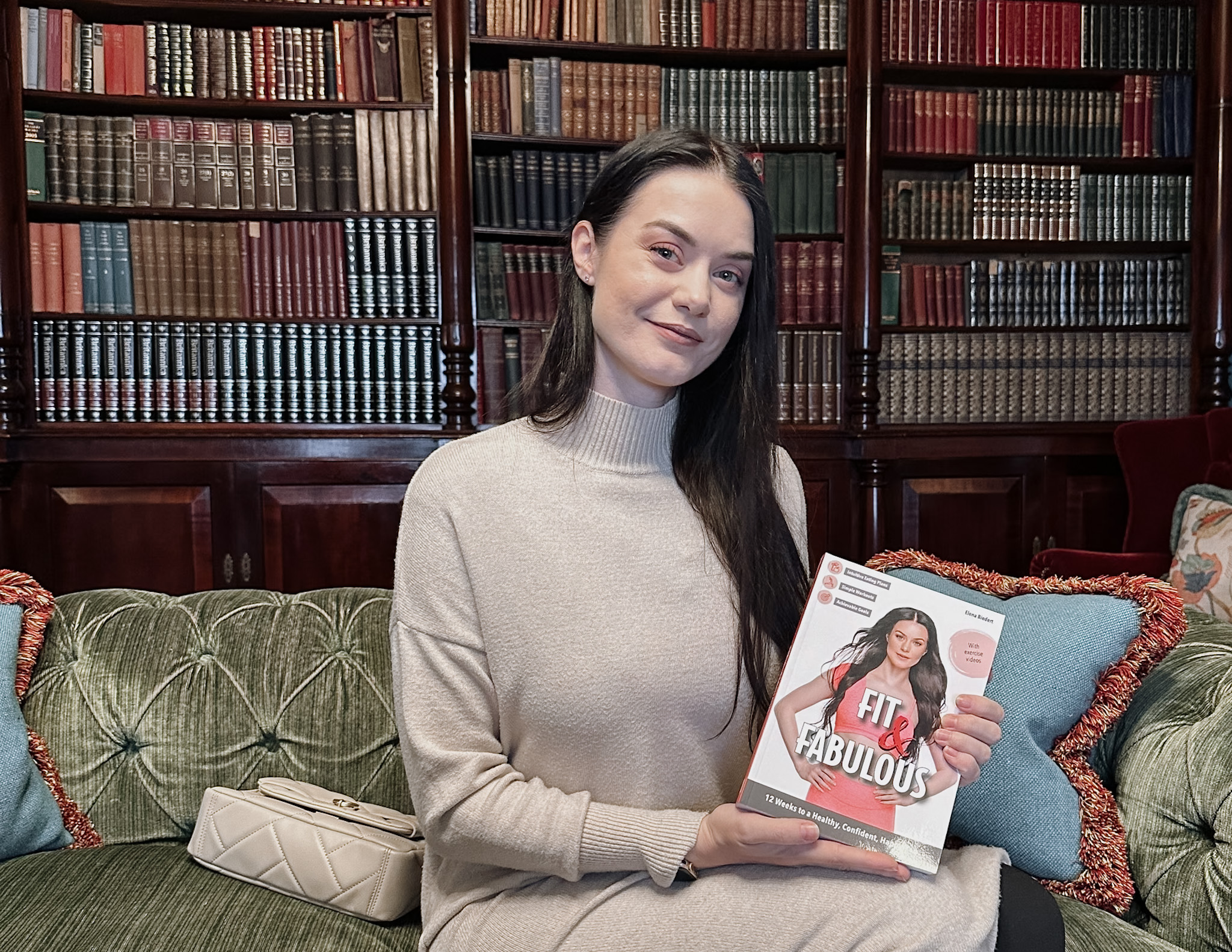 Elena Biedert sitting on the green couch in a library in London, holding her freshly released book Fit and Fabulous